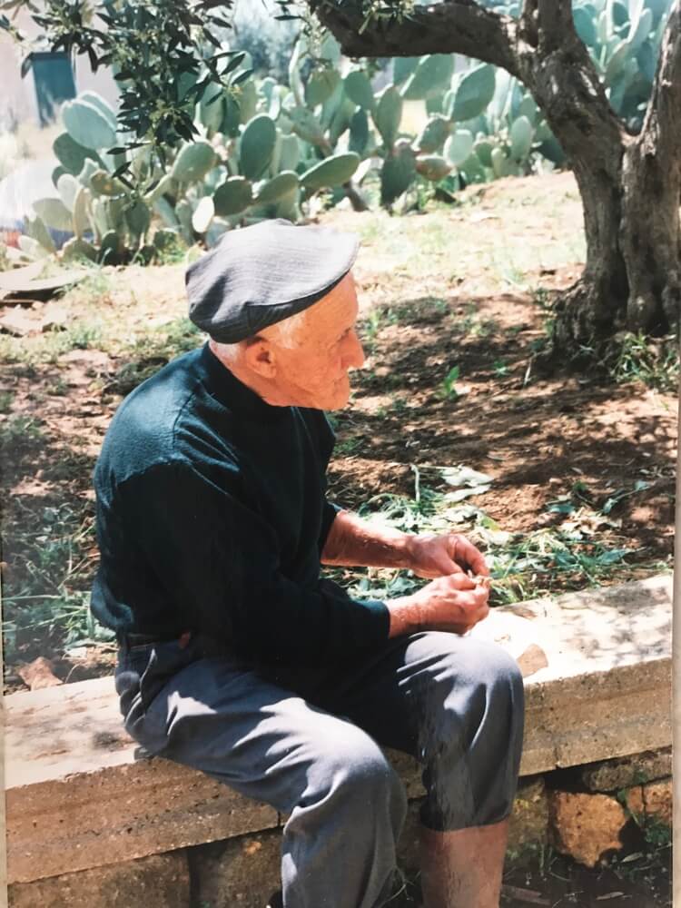 Uomo anziano seduto, berretto, mani in grembo, fichi d'india sullo sfondo.